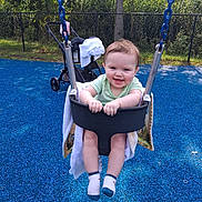 Bella is registered to the contest to win money with this photo: baby, child, swing, playground, smiling, happy, blue_surface, blanket, stroller, outdoor, trees, fence, person, feet, socks, seat, metal_chains, sunlight, greenery, fun
