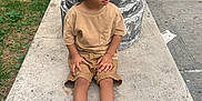 Abel is registered to the contest to win money with this photo: child, curly_hair, tan_clothing, white_sneakers, concrete, pillar, urban_park, greenery, side_view, sitting, thoughtful, outdoor, casual_wear, sidewalk, litter, daylight, young_boy, shorts, tshirt, park_bench
