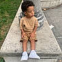 child, curly_hair, tan_clothing, white_sneakers, concrete, pillar, urban_park, greenery, side_view, sitting, thoughtful, outdoor, casual_wear, sidewalk, litter, daylight, young_boy, shorts, tshirt, park_bench