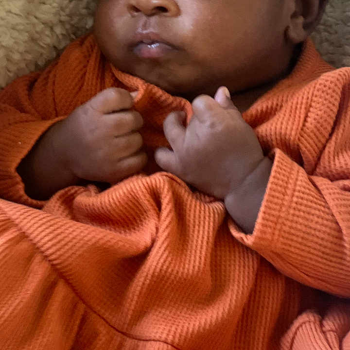 Nudai is registered to the contest to win money with this photo: baby, blanket, bow, child, clothing, cozy, cute, expression, face, fist, headband, indoors, infant, lying_down, newborn, orange_dress, portrait, skin, texture, young_child