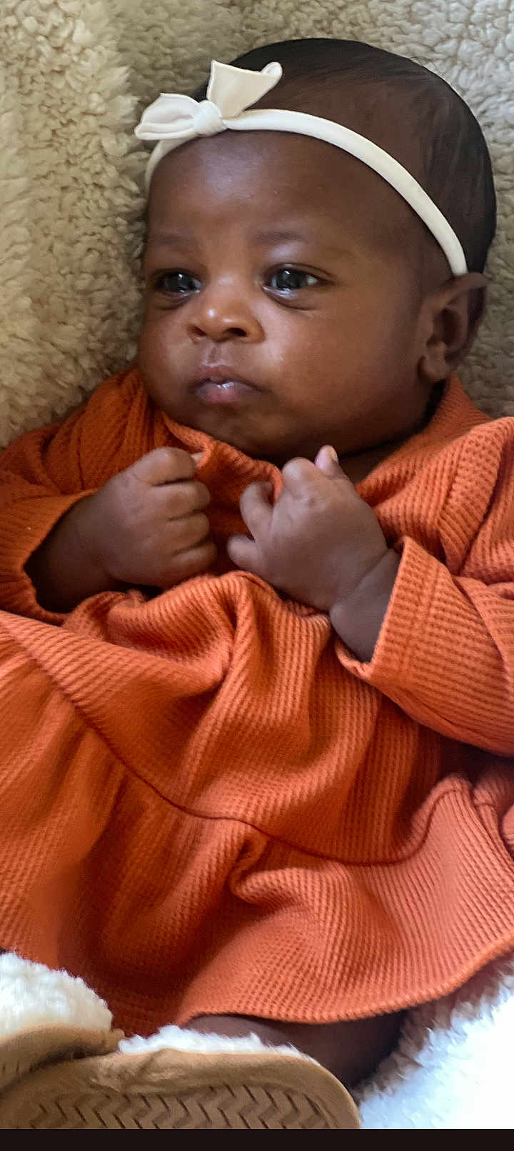 Nudai is registered to the contest to win money with this photo: baby, infant, face, headband, bow, orange_dress, clothing, fist, blanket, texture, skin, cute, portrait, young_child, newborn, expression, lying_down, indoors, cozy, child