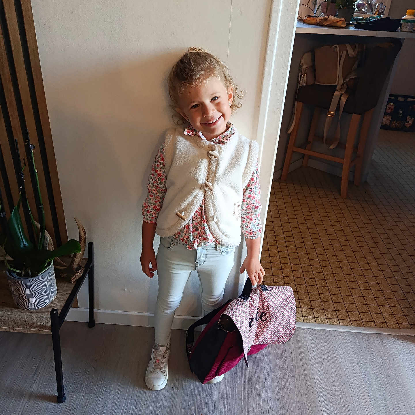 Lalie participe au concours pour gagner de l'argent avec cette photo : bag, chair, child, curly_hair, floor, floral_shirt, happy, home, indoor, kitchen, light_jeans, person, plant, smile, standing, table, wall, white_sneakers, white_vest, window