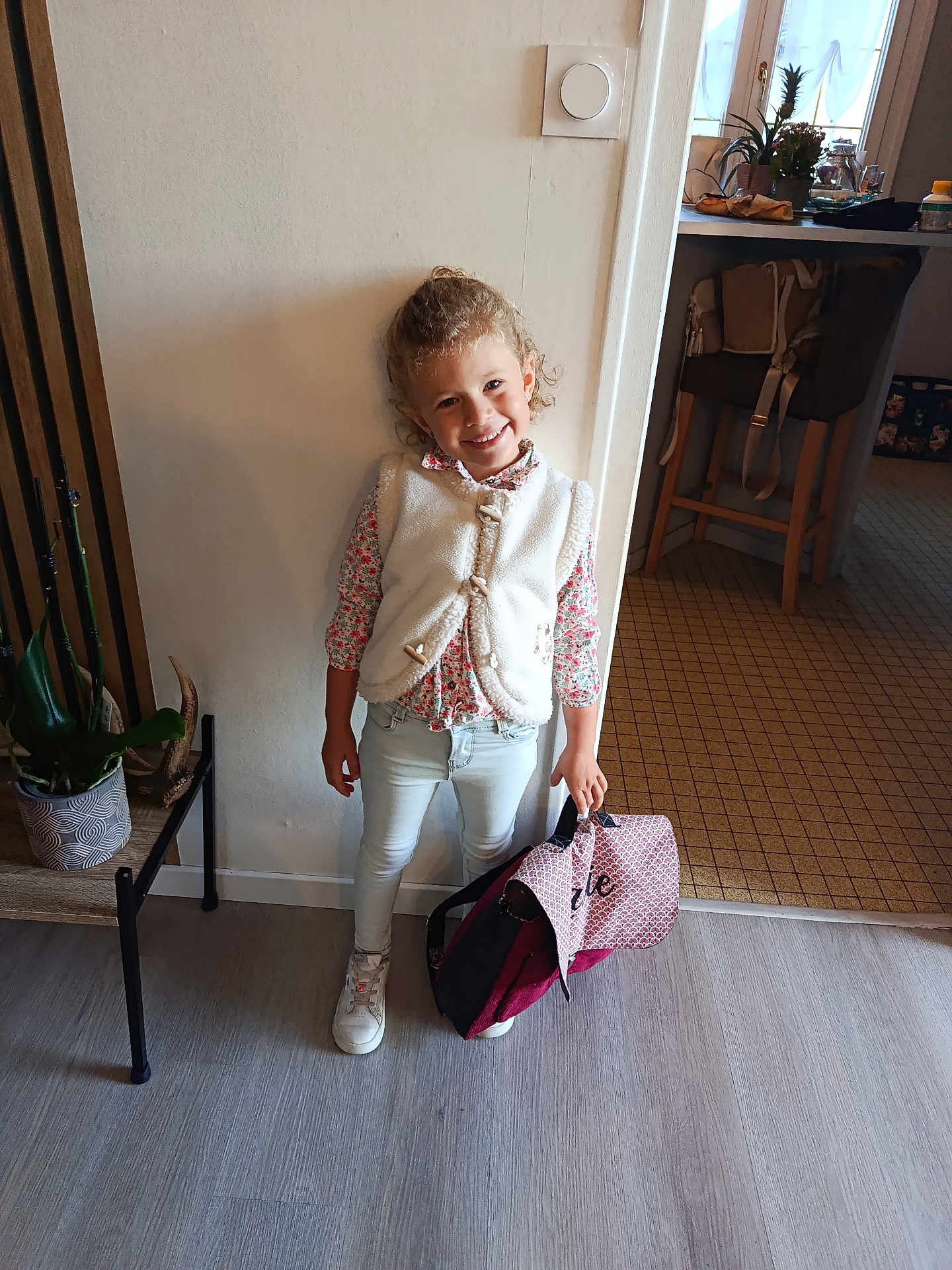 Lalie participe au concours pour gagner de l'argent avec cette photo : child, smile, curly_hair, indoor, wall, bag, white_vest, floral_shirt, light_jeans, white_sneakers, plant, chair, kitchen, floor, window, table, person, happy, standing, home
