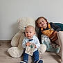 Lalie a rejoint le concours — aidez-le/la à gagner de superbes lots ! baby, child, teddy_bear, toy, floor, smile, overalls, card, curly_hair, blue_eyes, casual_clothing, indoor, wooden_floor, happy, cute, sitting, footwear, person, playful, family
