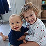 child, baby, smiling, happy, indoor, floor, curly_hair, blond_hair, blue_eyes, clothing, sitting, person, home, wooden_floor, cabinet, sock, cute, toddler, sibling, playful