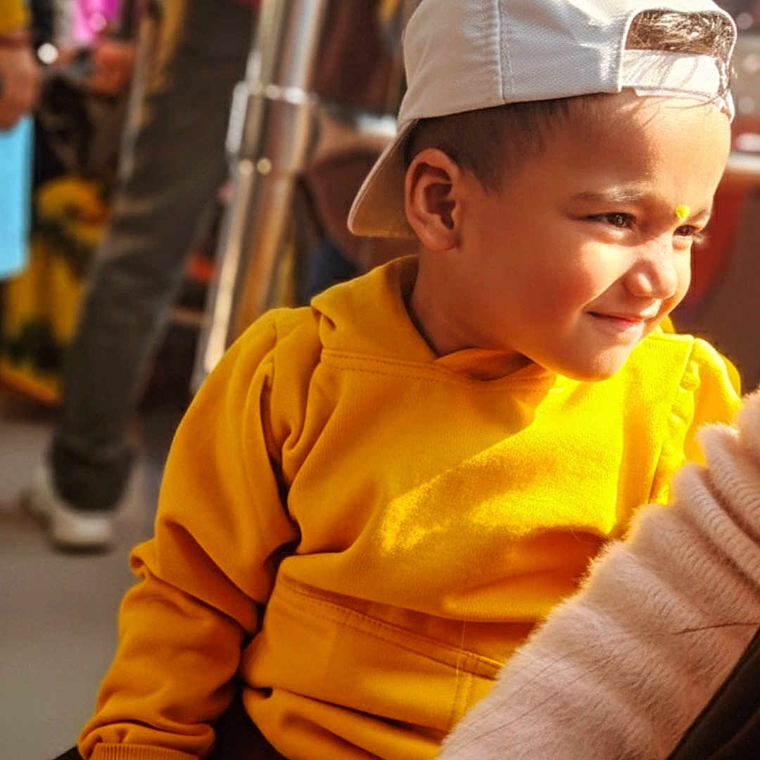Ansh joined the competition — help win amazing prizes! blurry_background, boy, candid, casual_clothing, child, close_up, fashion, hand, happy, indoor, people, person, portrait, seated, smiling, sunlight, warm_light, white_cap, yellow_hoodie, young