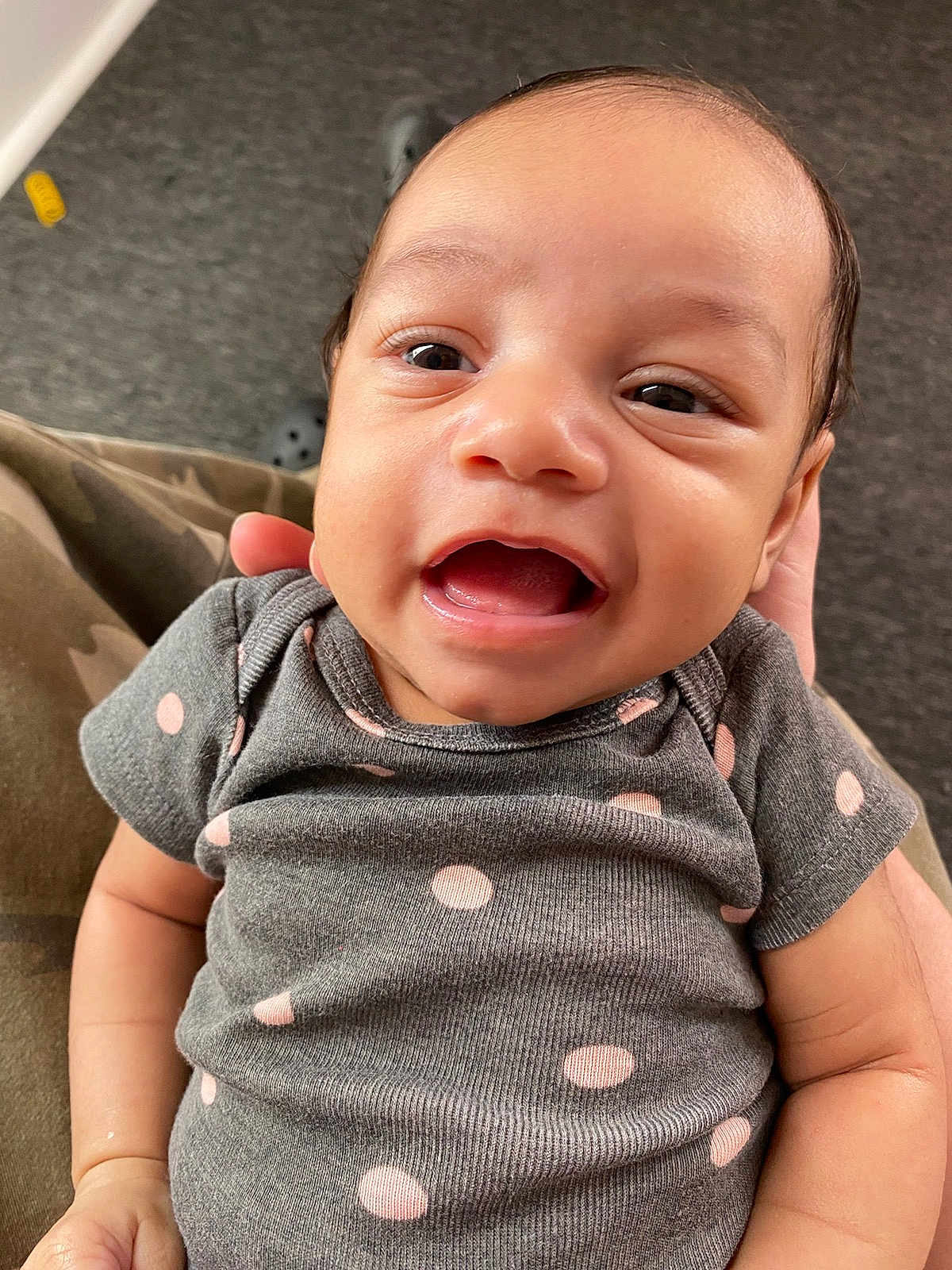 Rosalia joined the competition — help win amazing prizes! baby, child, smiling, face, onesie, polka_dot, grey_clothing, hand, carpet, toy, indoor, person, camouflage_pants, happy, cute, infant, portrait, closeup, skin, expression