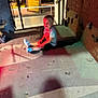 child, sitting, metal_floor, rivets, colorful_lighting, red_light, green_light, informational_sign, industrial, indoor, young_child, casual_clothing, shorts, tshirt, shoes, blurry, portrait, shadow, metal_surface, riveted_floor