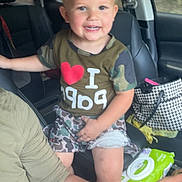 Gunner is registered to the contest to win money with this photo: child, toddler, smiling, car_interior, seat, tshirt, camo_clothing, handbag, baby_wipes, adult_arm, window, trees, seatbelt, shorts, person, happy, interior, travel, family, casual