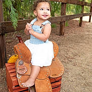 Zaya is registered to the contest to win money with this photo: toddler, child, smiling, outdoor, saddle, wooden_crate, denim, white_dress, curly_hair, fence, nature, greenery, happy, barefoot, portrait, rustic, playful, cute, sunlight, ground
