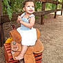 toddler, child, smiling, outdoor, saddle, wooden_crate, denim, white_dress, curly_hair, fence, nature, greenery, happy, barefoot, portrait, rustic, playful, cute, sunlight, ground