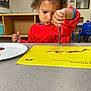 Zaya joined the competition — help win amazing prizes! child, toddler, painting, art, classroom, red_smock, curly_hair, yellow_paper, table, chair, education, kid, concentration, art_activity, indoor, person, young_child, preschool, learning, creative