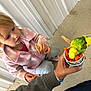 child, parakeet, bird, cup, carrots, seeds, hand, jacket, pink, green, yellow, floor, metal_wall, curious, feeding, interaction, person, casual_clothing, indoor, small_animal