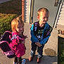 children, backpacks, sidewalk, sunlight, grass, shoes, jackets, smiling, standing, outdoor, building, door, concrete, happy, young, person, casual_clothing, daytime, portrait, kids