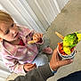 child, girl, bird, parakeet, feeding, cup, carrots, hand, pink_jacket, curious, indoor, floor, concrete, light_blue_pants, person, animal, pet, interaction, colorful, casual