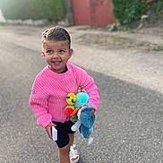 Lou participe au concours pour gagner de l'argent avec cette photo : child, girl, pink_sweater, toy, street, smile, happy, walking, outdoor, summer_clothing, sandals, shorts, daylight, playful, cute, person, hair_clip, nature, hedge, gate