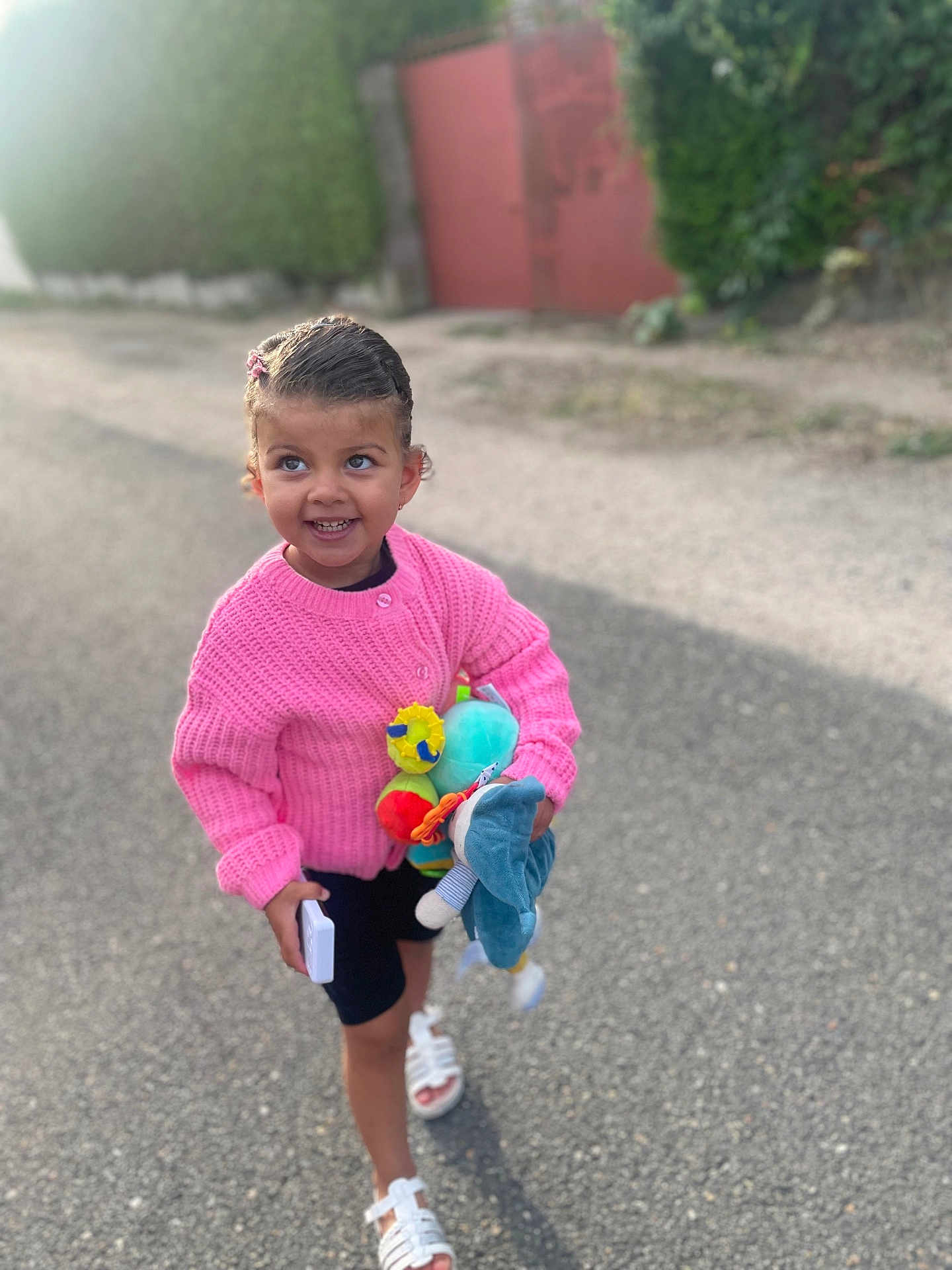 Lou participe au concours pour gagner de l'argent avec cette photo : child, girl, pink_sweater, toy, street, smile, happy, walking, outdoor, summer_clothing, sandals, shorts, daylight, playful, cute, person, hair_clip, nature, hedge, gate
