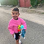 Lou participe au concours pour gagner de l'argent avec cette photo : child, girl, pink_sweater, toy, street, smile, happy, walking, outdoor, summer_clothing, sandals, shorts, daylight, playful, cute, person, hair_clip, nature, hedge, gate