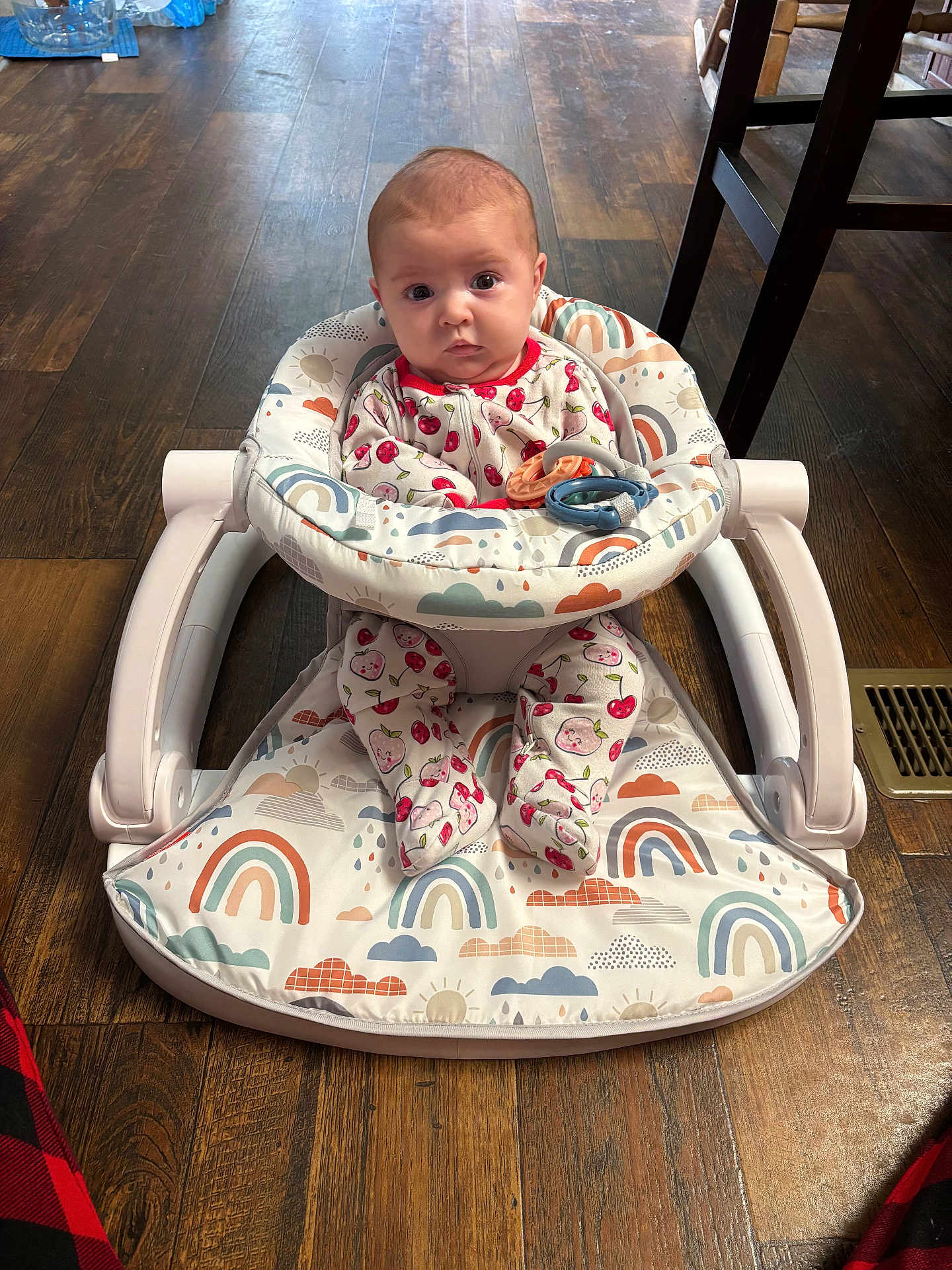 Sonney is registered to the contest to win money with this photo: baby, infant, baby_walker, rainbow_pattern, footwear, wooden_floor, indoor, clothing, face, person, sitting, toy, chair_leg, vent, cute, child, plush, patterned_clothing, expression, flooring
