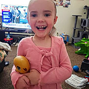 Oaklynn is registered to the contest to win money with this photo: child, smiling, pink_clothing, toy, living_room, television, carpet, furniture, cat_tree, ball, photo_wall, window, happy, indoor, person, playful, home, young_child, cute, holding