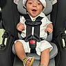 baby, infant, car_seat, smiling, hat, white_clothing, sandal, footwear, black_car_seat, child, happy, straps, safety_harness, indoors, cute, person, seat, headwear, face, young