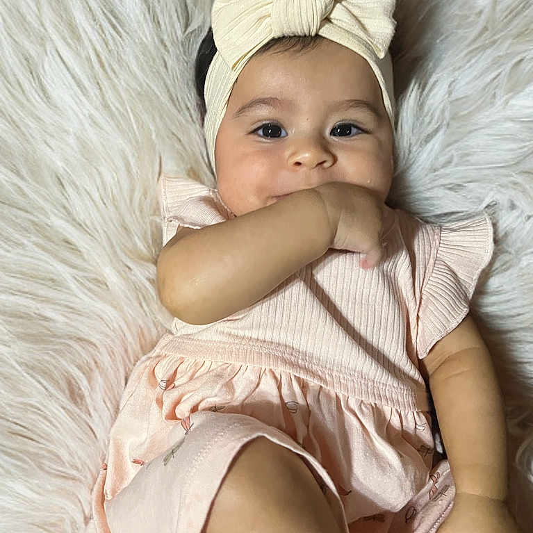 Eilany is registered to the contest to win money with this photo: baby, bow, child, close_up, comfortable, cute, eyes, face, fluffy_rug, girl, hand_in_mouth, headband, indoor, infant, pink_dress, portrait, skin, smiling, soft_texture, young