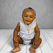 Hilton is registered to the contest to win money with this photo: baby, smiling, sitting, floor, wooden_floor, gray_wall, portrait, child, cute, indoors, necklace, clothing, happy, infant, person, studio, casual, cute_baby, one_person, fashion