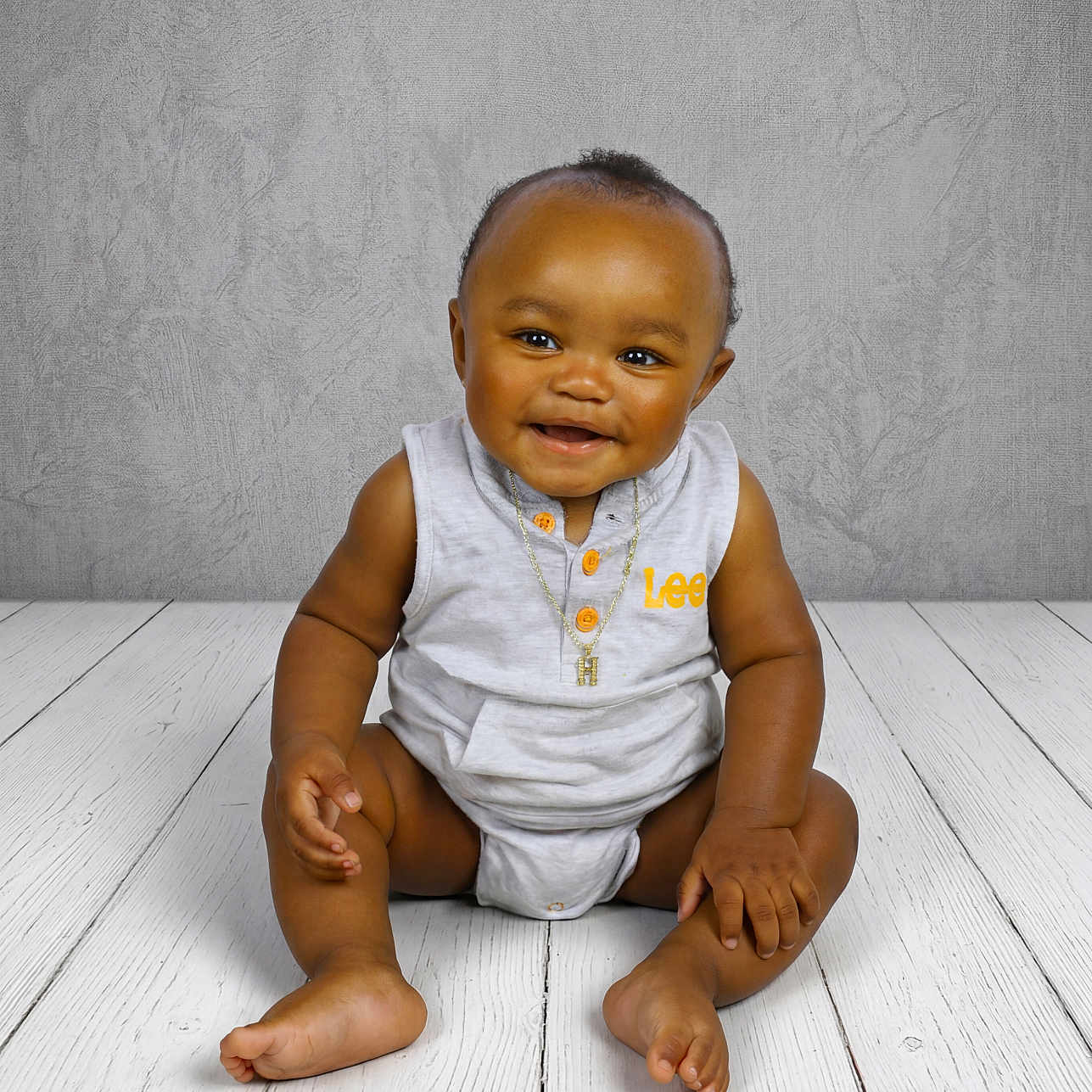 Hilton is registered to the contest to win money with this photo: baby, smiling, sitting, floor, wooden_floor, gray_wall, portrait, child, cute, indoors, necklace, clothing, happy, infant, person, studio, casual, cute_baby, one_person, fashion