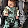 baby, blanket, car_seat, eyes_open, fabric, face, green_overalls, indoor, infant, looking_away, newborn, onesie, overalls, portrait, seat_padding, soft_lighting, tiny_feet, tiny_hands, white_shoes, wood_floor
