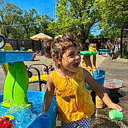 Domila joined the competition — help win amazing prizes! child, toddler, girl, outdoor, playground, water_play, splash_table, swimsuit, yellow_shirt, patterned_pants, green_cup, smiling, laughing, summer, sunlight, trees, backyard, pool, toys, portrait