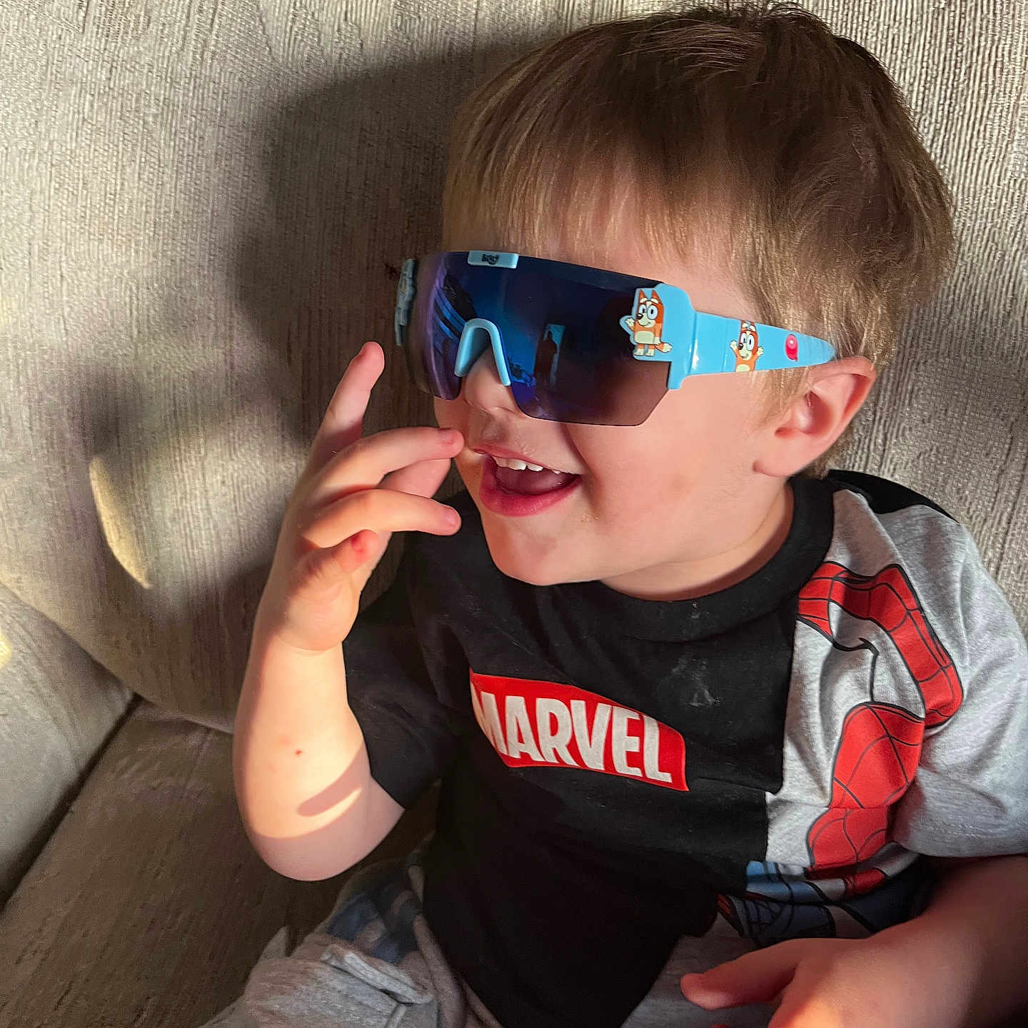 Leelend is registered to the contest to win money with this photo: child, boy, sunglasses, marvel, tshirt, smiling, laughing, hand, sofa, couch, indoor, casual_clothing, short_hair, light_shadow, playful, portrait, relaxed, furniture, young, happy