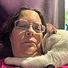 woman, cat, white_cat, pet, cuddling, glasses, face, portrait, selfie, indoor, window, sunlight, purple_wall, pink_clothing, smartphone, pendant, fluffy, relaxed, closeup, companionship