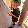 child, boy, black_tshirt, cap, hair_clipper, standing, balcony, railing, outdoor, concrete_floor, shorts, black_shoes, curious, looking_up, daylight, urban, person, casual, young, portrait