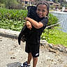 child, puppy, black_dog, outdoor, smiling, braids, nike, shorts, sneakers, water, greenery, daylight, happy, person, nature, pet, holding, portrait, casual_clothing, playful