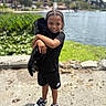 child, dog, hugging, lake, outdoor, greenery, water, smiling, black_clothing, shorts, sneakers, sunny, nature, pet, young_person, casual_wear, portrait, happy, daytime, park