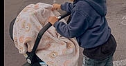 Ak is registered to the contest to win money with this photo: child, stroller, blanket, parking_lot, cap, hoodie, shorts, sneakers, smiling, outdoor, urban, sidewalk, baby, toy, playful, person, vehicle, wheel, road, daytime