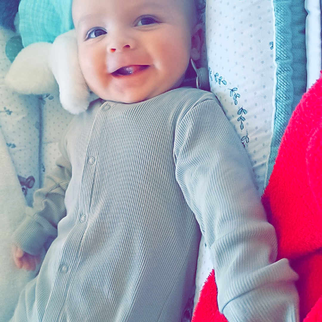 Timothée participe au concours pour gagner de l'argent avec cette photo : baby, baby_nest, bed, blanket, child, comfort, cozy, cute, face, happy, indoor, infant, lying_down, onesie, patterned, portrait, red_blanket, smiling, soft_toy, toy