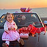 child, toddler, car, red_bow, sunset, ocean, pink_dress, smiling, curly_hair, outdoor, happy, toy_purse, balloons, reflection, vehicle, holiday, celebration, cute, fashion, portrait