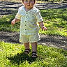 toddler, child, grass, outdoor, sunlight, shorts, shirt, sandals, face, person, nature, trees, furniture, wood, yard, daylight, curious, standing, summer, play