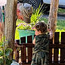 adult, camouflage_clothing, cap, child, curly_hair, daylight, garden, grass, hat, interaction, man, outdoor, plant, potted_plant, repair, toddler, tool, vehicle, wheel, wooden_fence