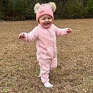 Serenity is registered to the contest to win money with this photo: toddler, child, pink_hat, pink_outfit, smiling, walking, grass, outdoor, trees, winter_clothing, happy, nature, baby, cute, fur_pom_poms, one_person, headwear, full_body, cold_weather, playful