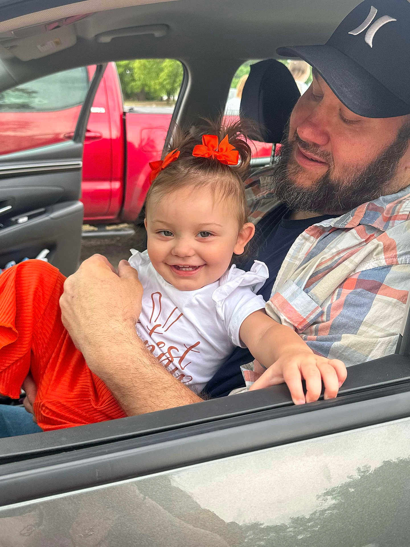 Serenity is registered to the contest to win money with this photo: toddler, child, man, car, window, seat, smile, hair_bow, orange_clothing, cap, beard, plaid_shirt, hand, red_truck, outdoor, greenery, happy, family, portrait, casual