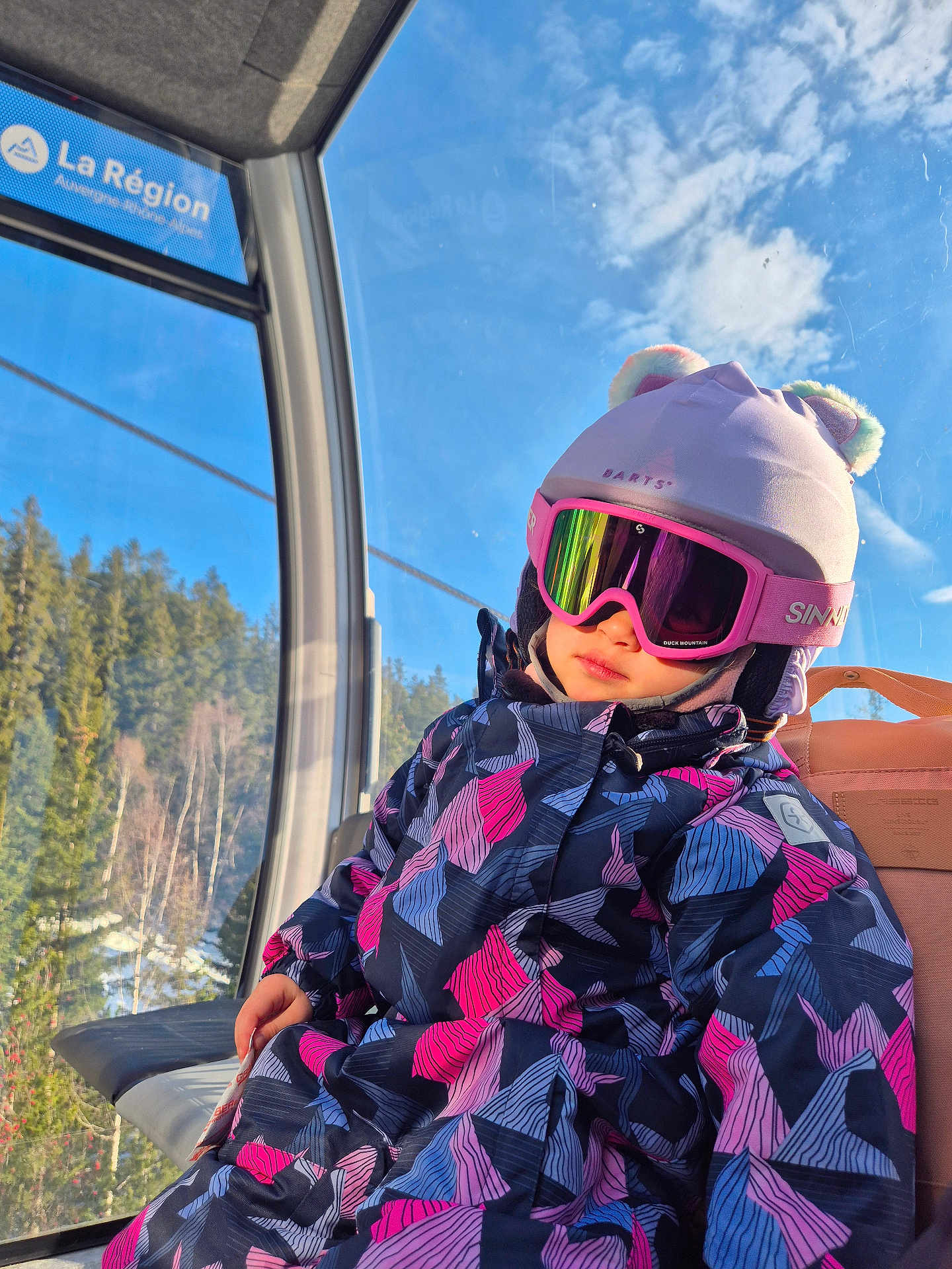 Enola a rejoint le concours — aidez-le/la à gagner de superbes lots ! child, ski_goggles, winter_clothing, hat, playful_hat, gondola, cabin, mountain, forest, sky, winter, snow, outdoor, reflection, colorful_jacket, pink, blue, seat, scenery, sunlight