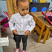 Enola a rejoint le concours — aidez-le/la à gagner de superbes lots ! child, girl, indoor, white_shirt, leggings, pink_sneakers, name_tag, toy_blocks, doll, table, floor_tiles, person, playroom, furniture, child_play, casual_clothing, standing, young_child, curly_hair, serious_expression