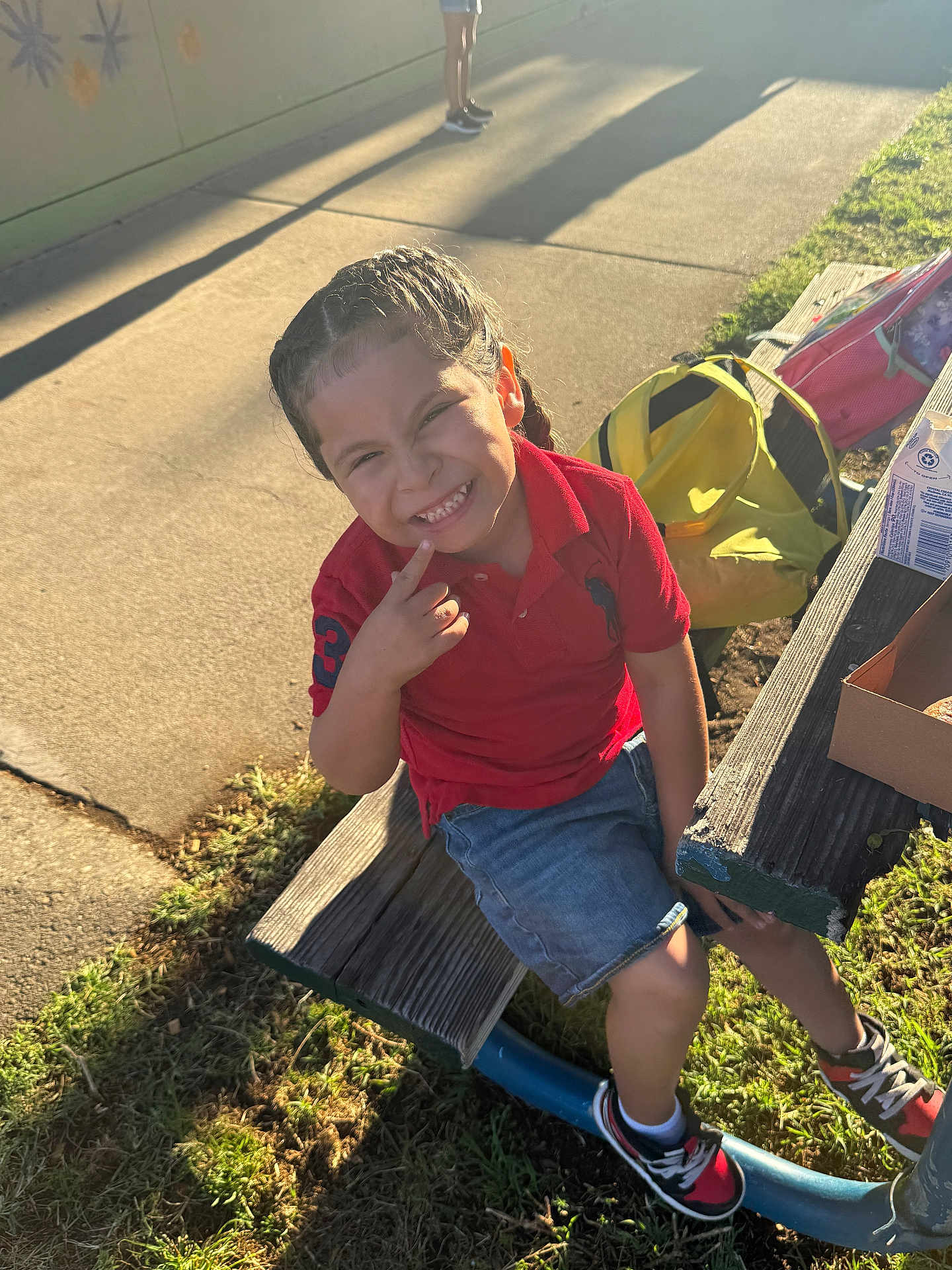 Mateo joined the competition — help win amazing prizes! child, smile, outdoor, bench, sunlight, shadow, grass, pavement, braided_hair, red_shirt, denim_shorts, sneakers, backpack, carton, happy, person, casual_clothing, playground, daylight, portrait