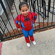 Mateo is registered to the contest to win money with this photo: blue, boy, child, confident, costume, daylight, fence, hair, outdoor, person, playful, red, sidewalk, smile, spiderman_costume, standing, superhero, urban, white_shoes, young