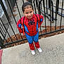 blue, boy, child, confident, costume, daylight, fence, hair, outdoor, person, playful, red, sidewalk, smile, spiderman_costume, standing, superhero, urban, white_shoes, young