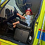 child, boy, firefighter_hat, smiling, steering_wheel, fire_truck, vehicle_interior, seat, black_shoes, shorts, tshirt, window, door, dashboard, pedals, sunlight, outdoor, yellow, red, happy