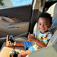 Donyè is registered to the contest to win money with this photo: child, toddler, car_interior, seatbelt, smiling, sneakers, mickey_mouse, shirt, denim_shorts, sunlight, window, sky, clouds, door_panel, car_seat, happy, person, seat, sitting, casual_clothing