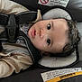 baby, car_seat, harness, child, infant, face, curly_hair, eyes, clothing, warning_label, seat, straps, head_support, indoor, closeup, expression, cute, young, sitting, person